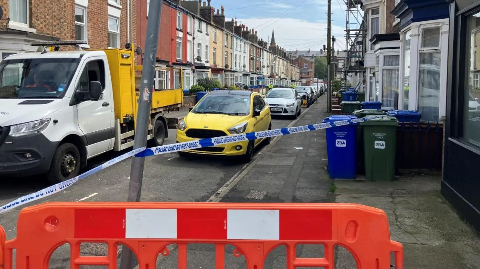 The image shows a residential street lined with terraced houses. The street has been partially cordoned off. At the bottom of the image is a bright orange plastic road barrier, positioned across the pavement as a physical block. Just behind it, blue-and-white police tape stretches across the street and pavement. The tape reads POLICE LINE DO NOT CROSS. A yellow car is parked on the left side of the street, just behind the police tape. In front of it is a white flatbed work truck. The street is lined with closely parked cars extending into the distance on both sides.
