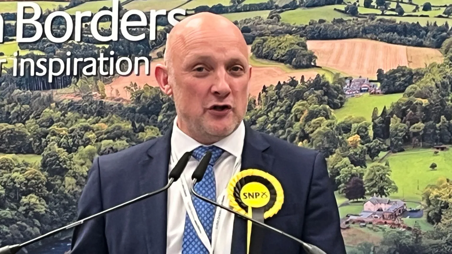 Callum Kerr - a bald man in a suit with an SNP rosette - stands in front of photograph of the countryside, speaking into two microphones. 