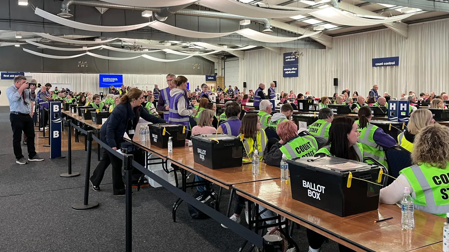 An election count with numerous ballot boxes and people in yellow high visibility tops in a large hall