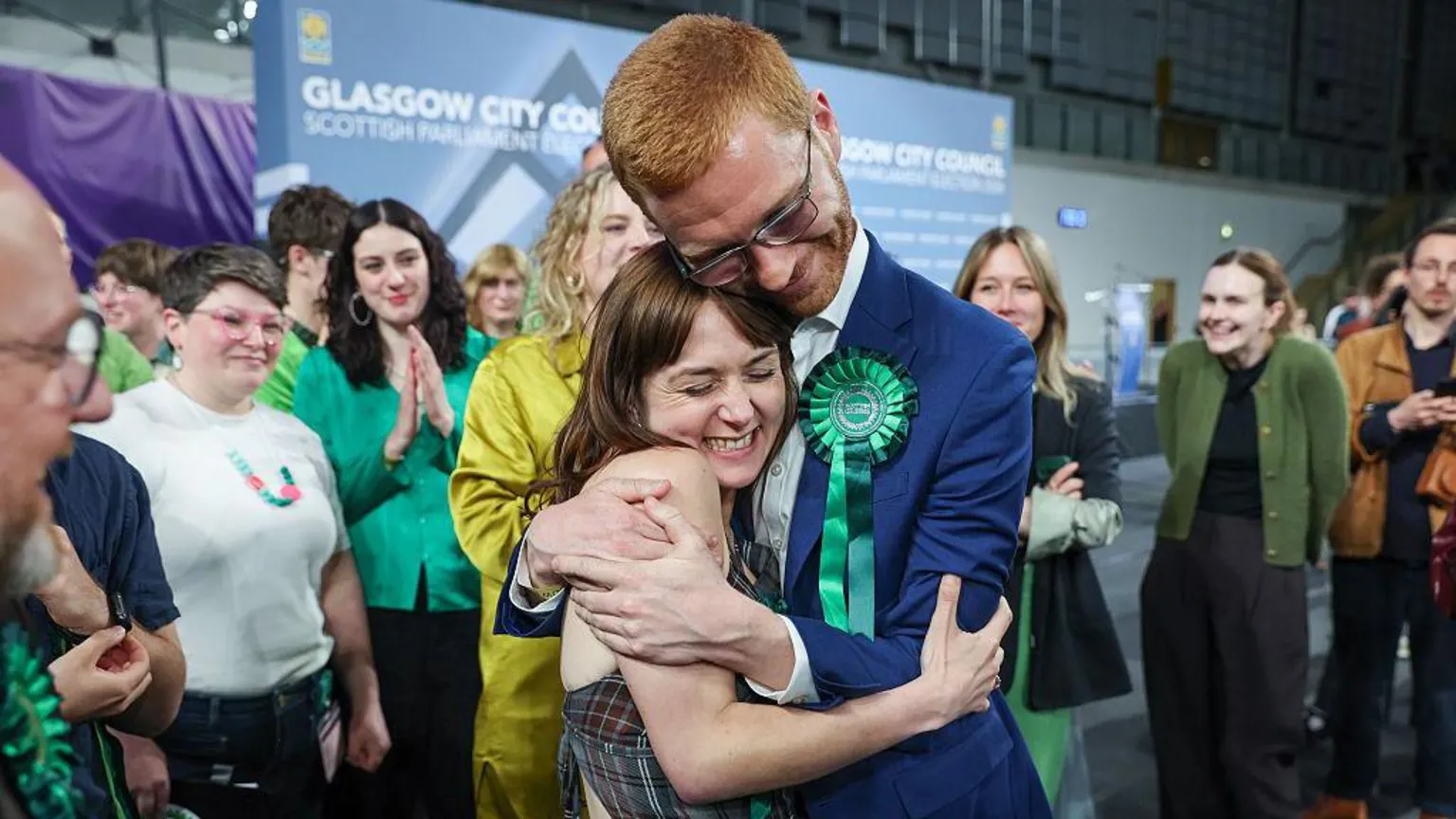  Holly Bruce - a woman in a patterned dress and and with brown hair, is hugged by Ross Greer, a man in a bright blue suit with green rosette, while smiling Green party volunteers look on. 