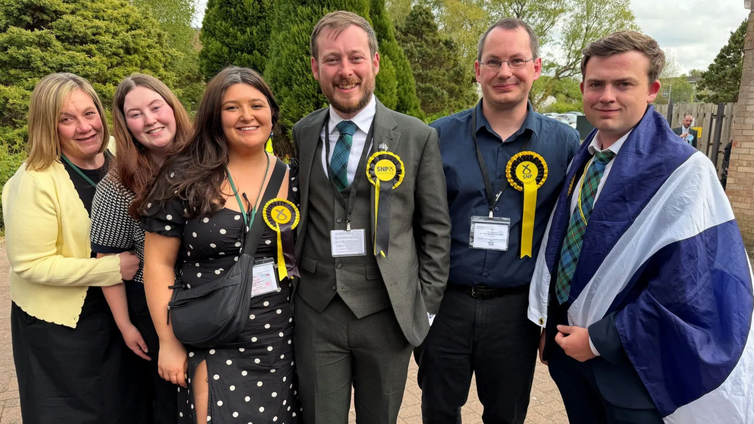 A row of six people - three women and three men - smiling and celebrating after election victory. Most of them are wearing SNP rosettes, one has a Saltire wrapped around them. Alex Kerr is in the centre - a man with a suit and a beard.