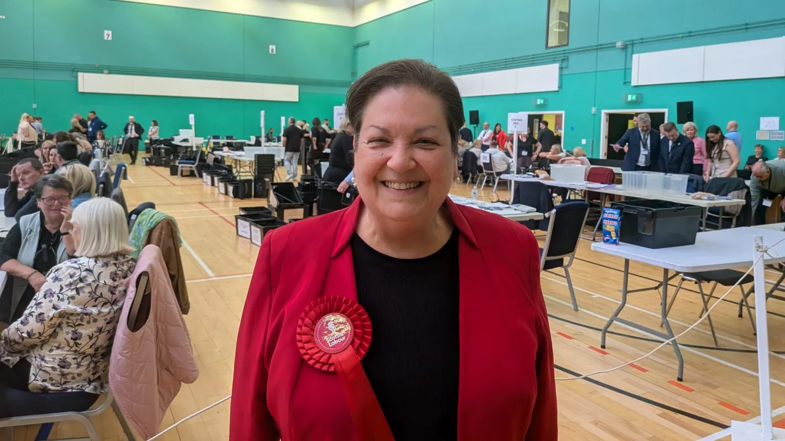 Jackie Baillie - a woman with a red jacket and dark hair, on the floor of an election count. She is smiling for the camera.