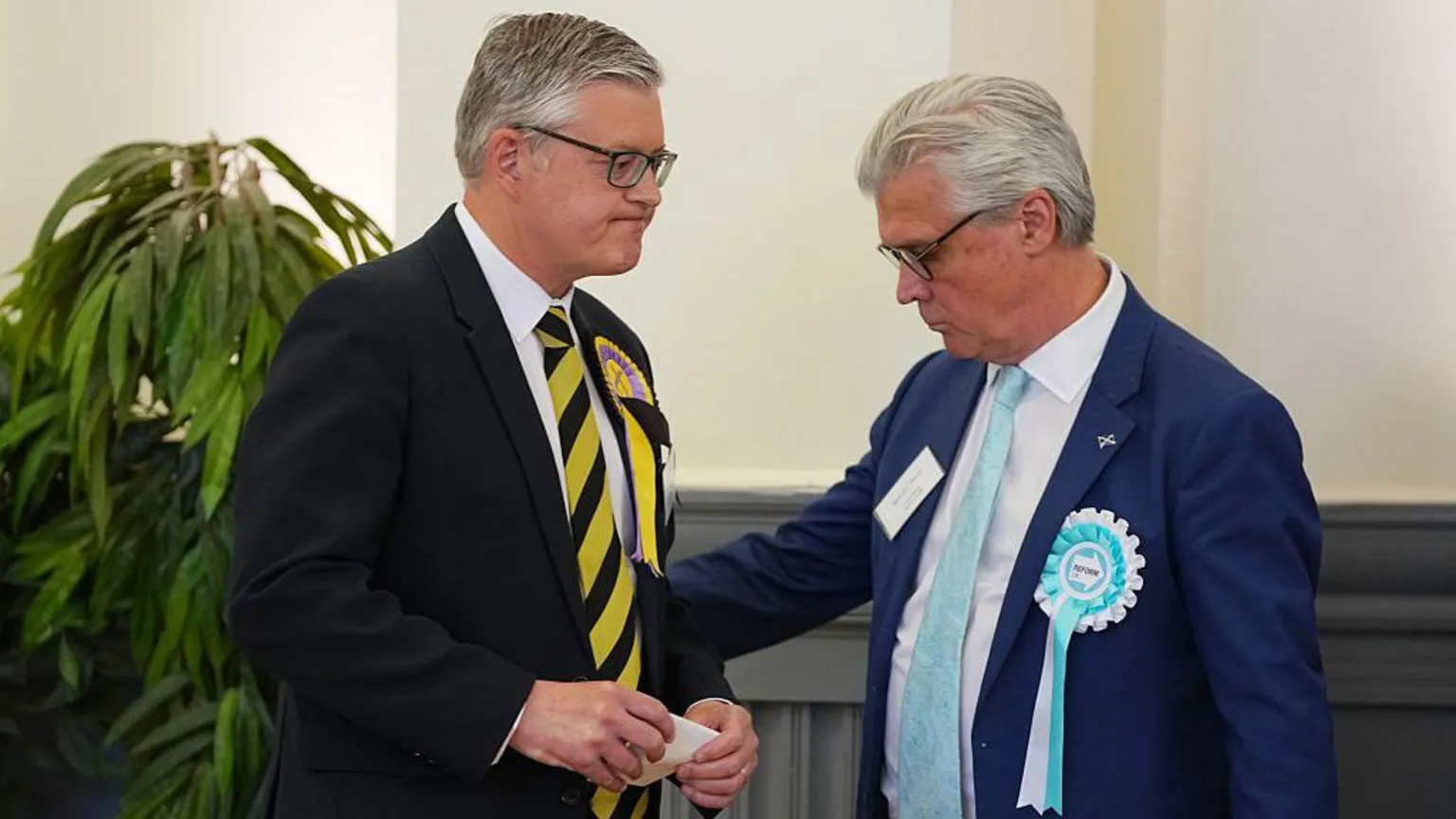 Stuart McMillan, wearing a dark suit and yellow tie, heads towards a podium to make a victory speech. Malcolm Offord, a man with silver hair and wearing a blue suit, pats his arm as he goes by.