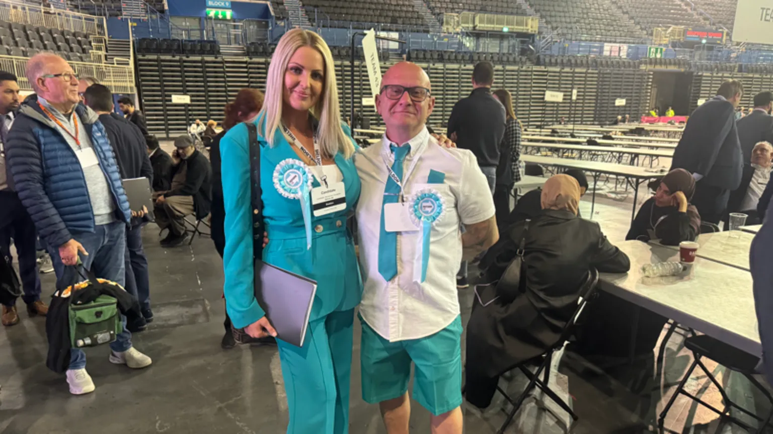 A woman with blonde hair and a bald man stand next to each other and smile at the camera. They both wear Reform UK's party colours of light blue and white, as well as rosettes.