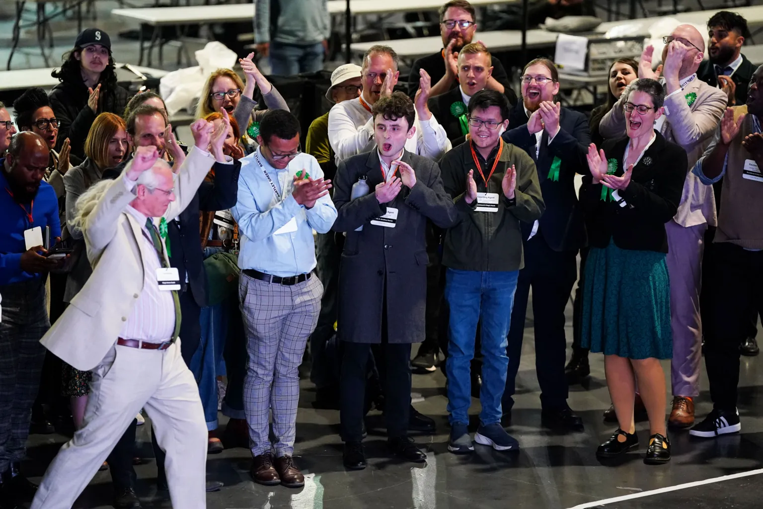 Jacob King/PA Wire A man in a cream suit with long white hair holds his arms aloft as a group of people wearing green rosettes cheer behind him 