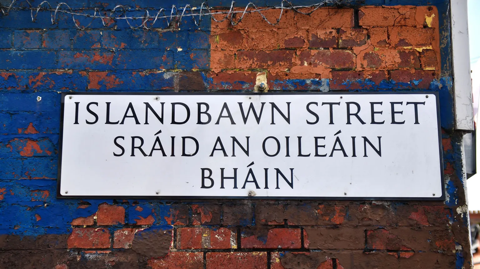  A white bilingual street sign in Irish and English. It is on a red brick wall that is partially painted blue. Some barbed wire can be seen on the wall above the sign. 