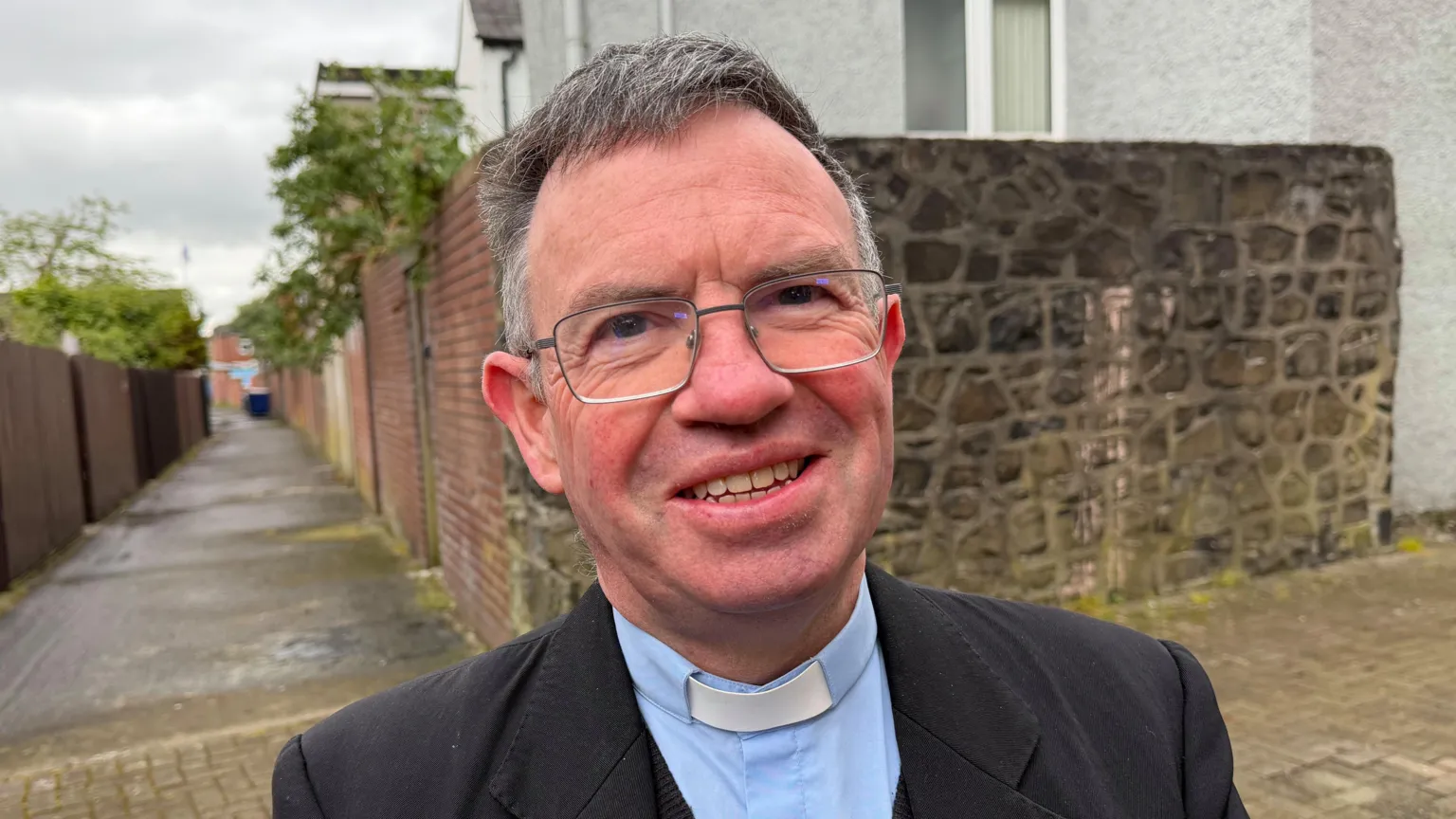A man smiles at the camera. He has large, thin rimmed glasses and wears a priest's clothes - blazer, shirt, clerical collar. 