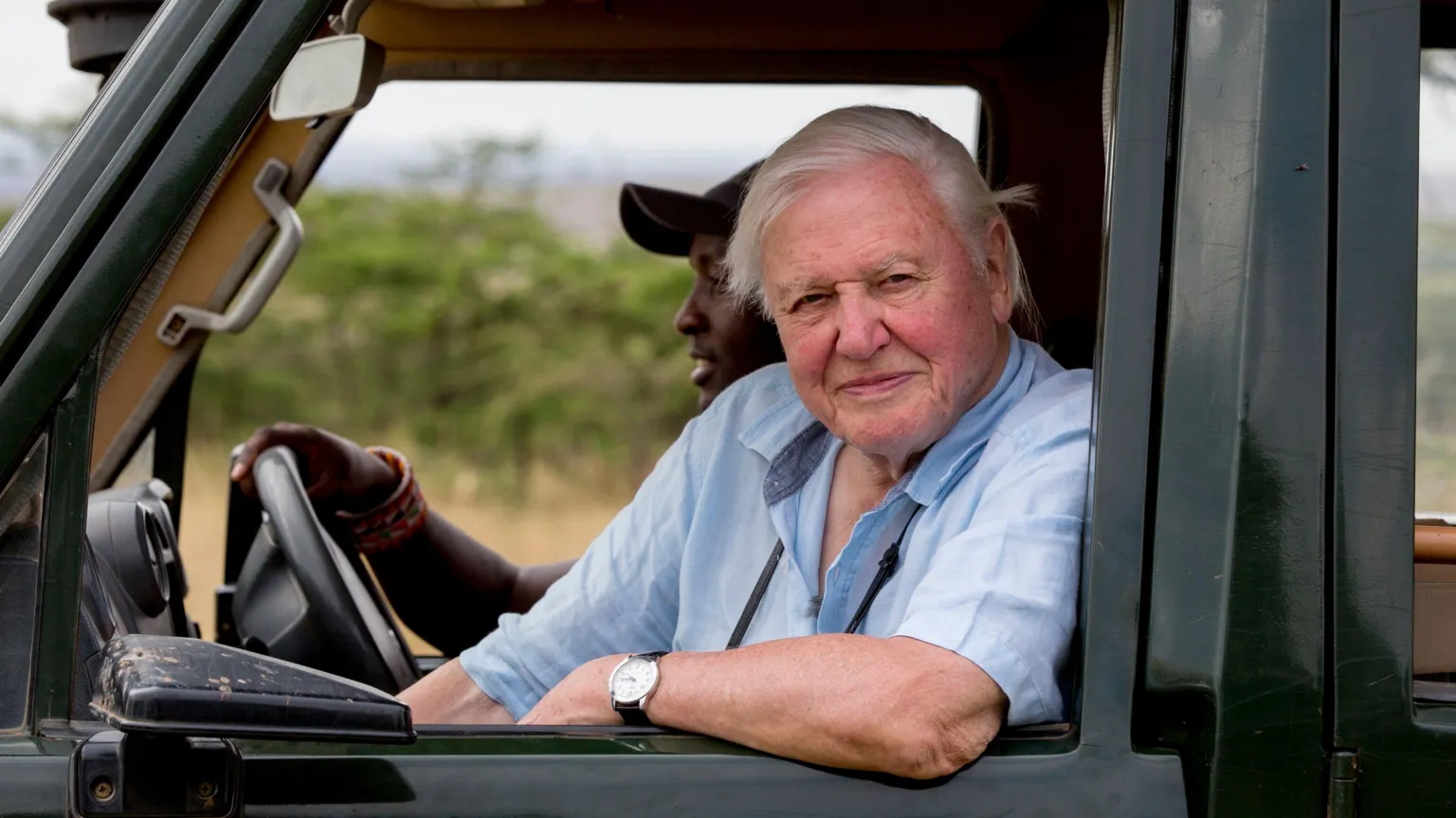 Silverback Films Sir David Attenborough is looking out of the window of a 4 by 4 which is being driven by a man in a cap. Sir David is wearing a pale blue short-sleeve shirt and has his arm on the car door. He is smiling at the camera.