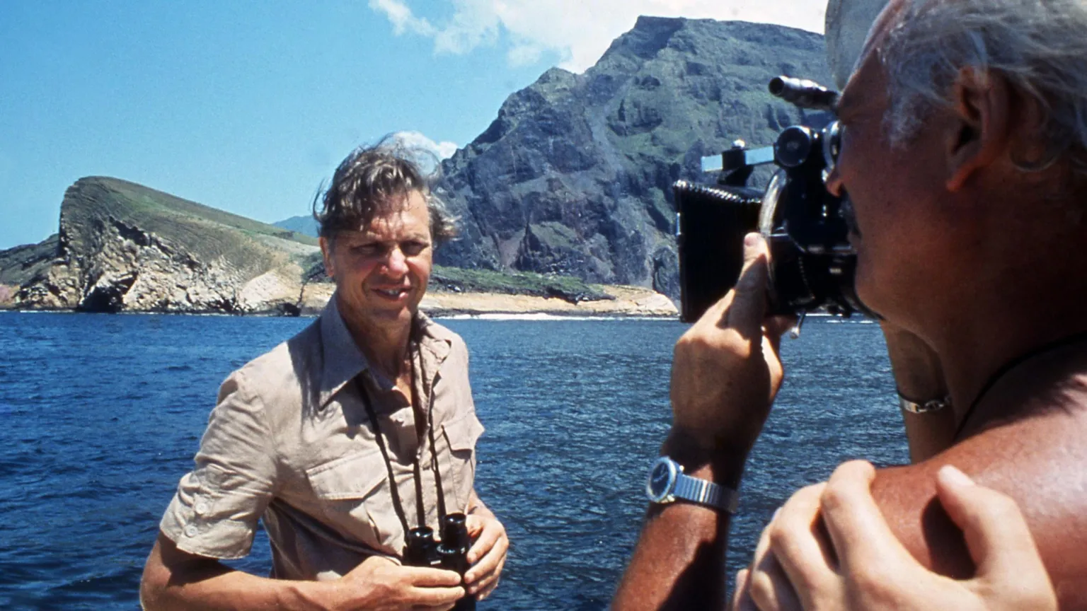 David Attenborough pictured in 1979 in a light brown camo shirt. He is standing in front of a coastline with the sea behind him, being filmed by a camera man. He is holding a pair of binoculars round his neck.