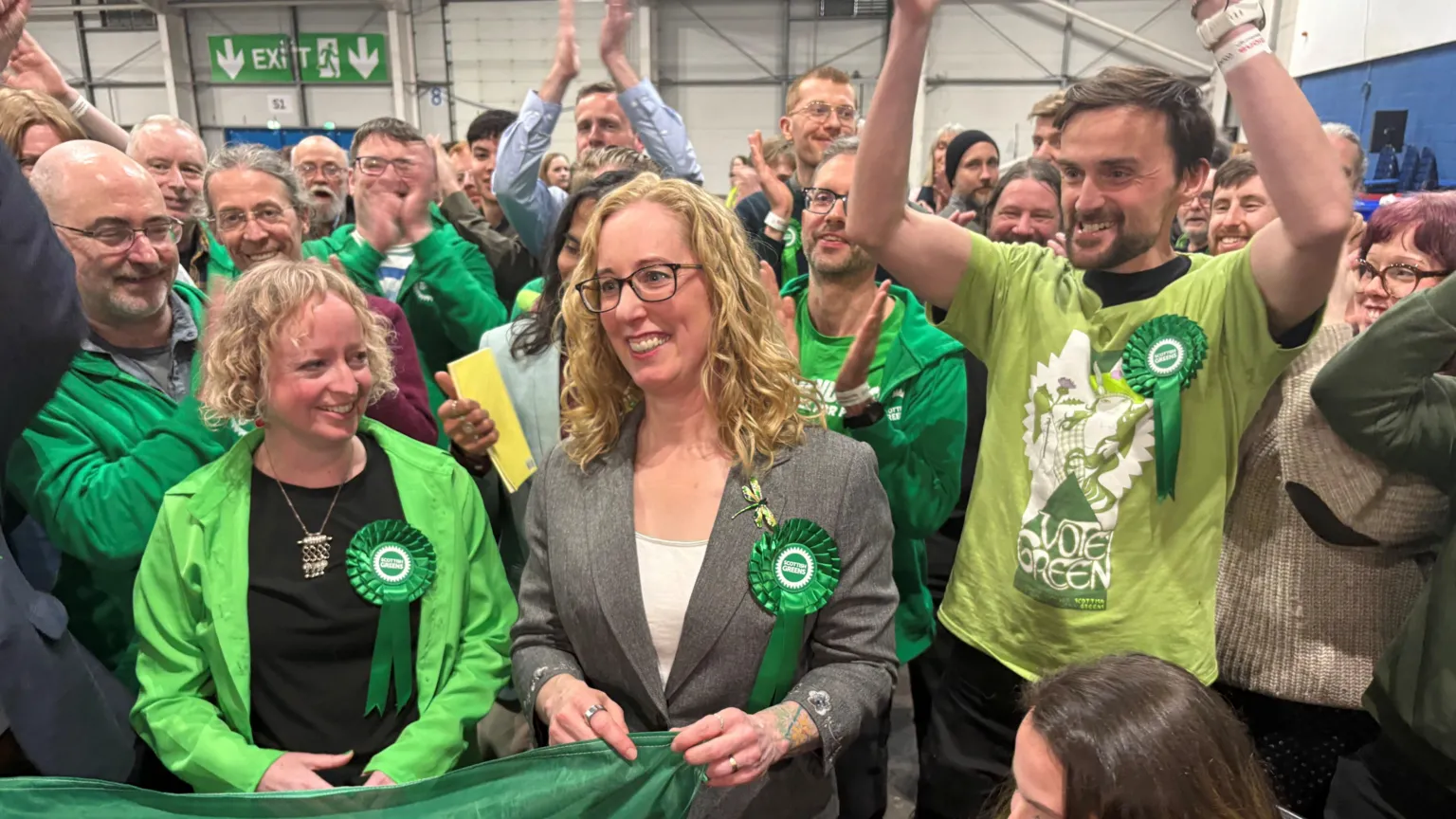  Lorna Slater, in grey suit and white shirt, is surrounded by Green Party supporters, largely in green. They look very happy. They are in a modern industrial-looking hall.