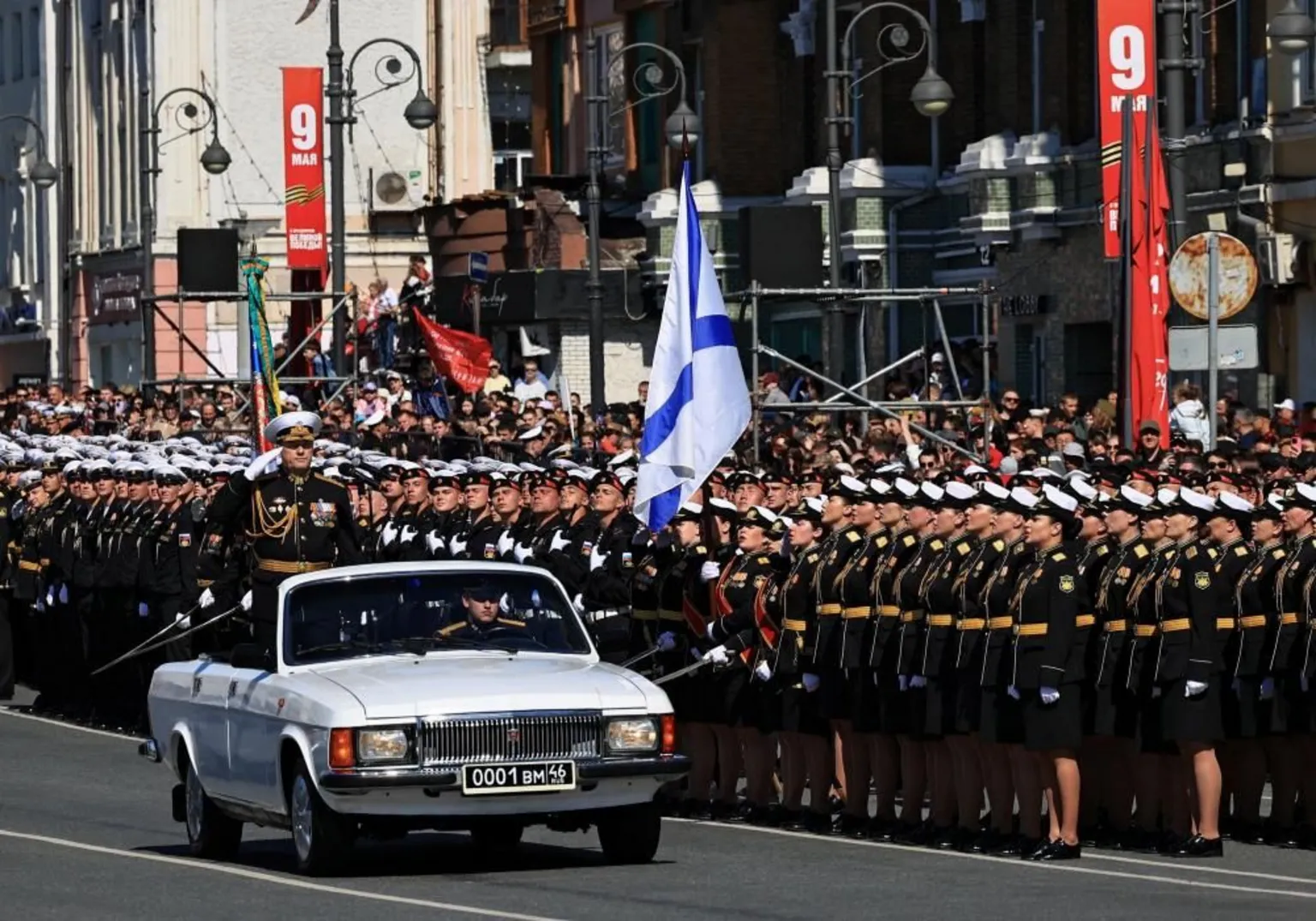  A Russian military commander salutes while standing in an open-top, white car being driven by another soldier on a road flanked by rows of military personnel. 