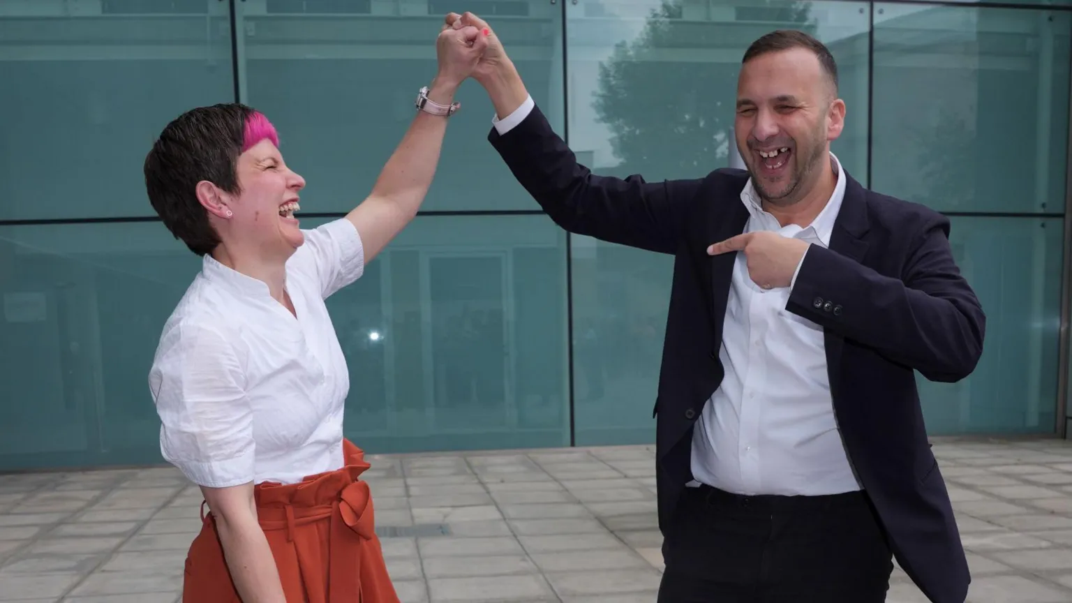  Zoë Garbett, left, seen from side view, laughs, smiles and does a high hand-shake with Jack Zack Polanski as they celebrate her mayoral win. She has short hair which is dark apart from a pink fringe and wears a white blouse and red skirt. He wears dark suit and white shirt.