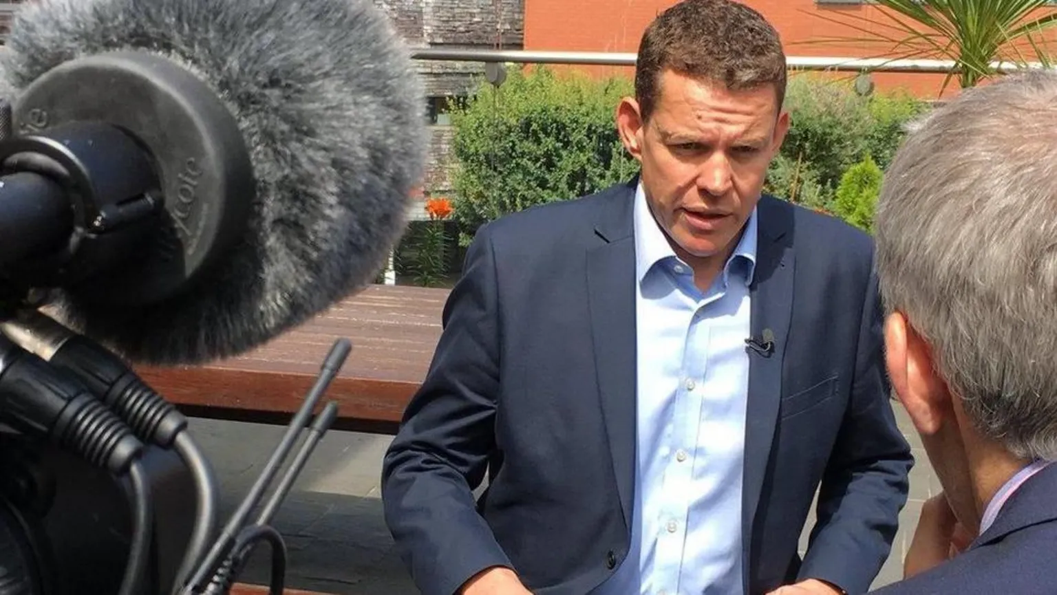 Rhun ap Iorwerth in sunlight being questioned by a BBC reporter with a camera microphone visible on the left of the picture.