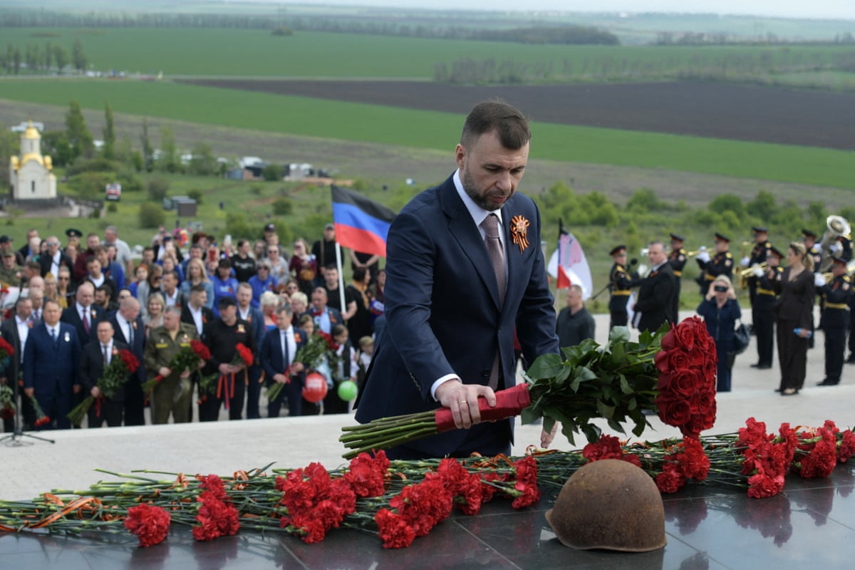 Denis Pushilin is facing the camera as he lays down flowers at the Savur-Mohyla memorial. Behind him is a small crowd and green fields. 