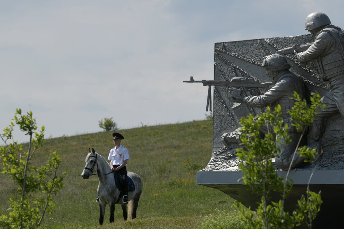 A uniformed Russian police officer sits on a white and grey horse next to a monument showing soldiers holding guns. 