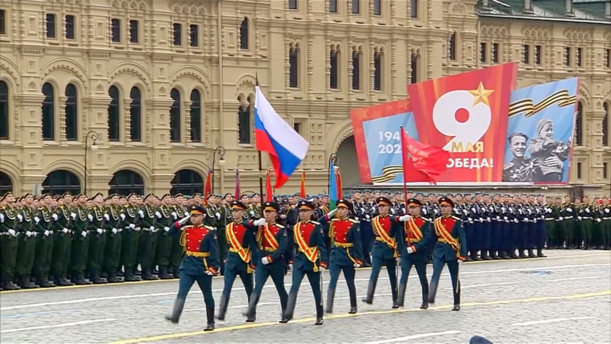 Eight uniformed Russian military service members march while holding a Russian flag overhead down a path in front of several tight lines of other uniformed Russian service members in Moscow's Red Square. 