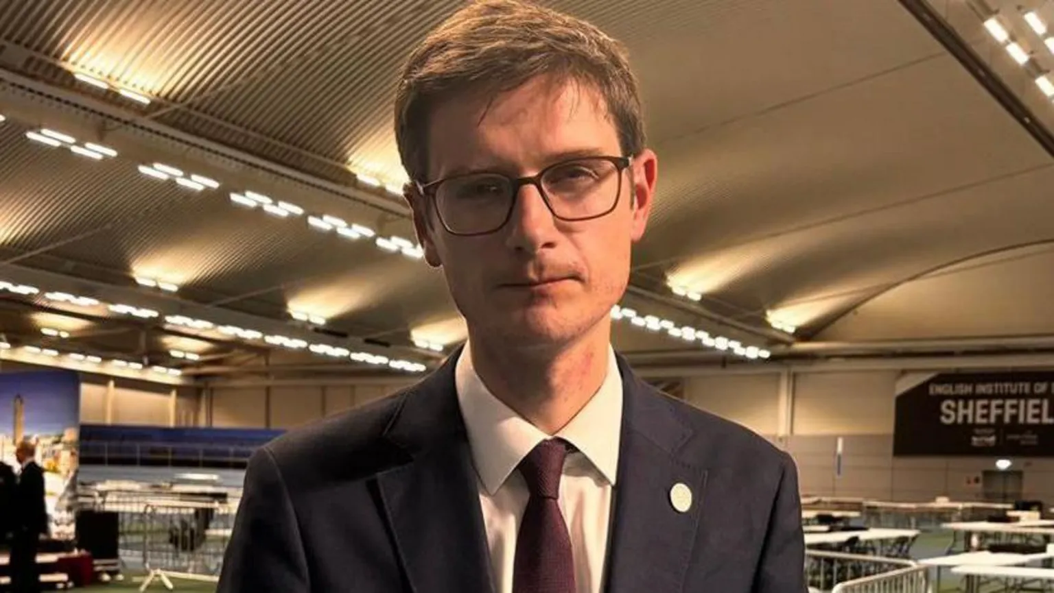 Simon Thake/BBC A man stands in a leisure centre where tables are in the background for the vote count. He has a navy suit, burgundy tie, brown short hair and black square glasses. He has a neutral expression but looks sad.