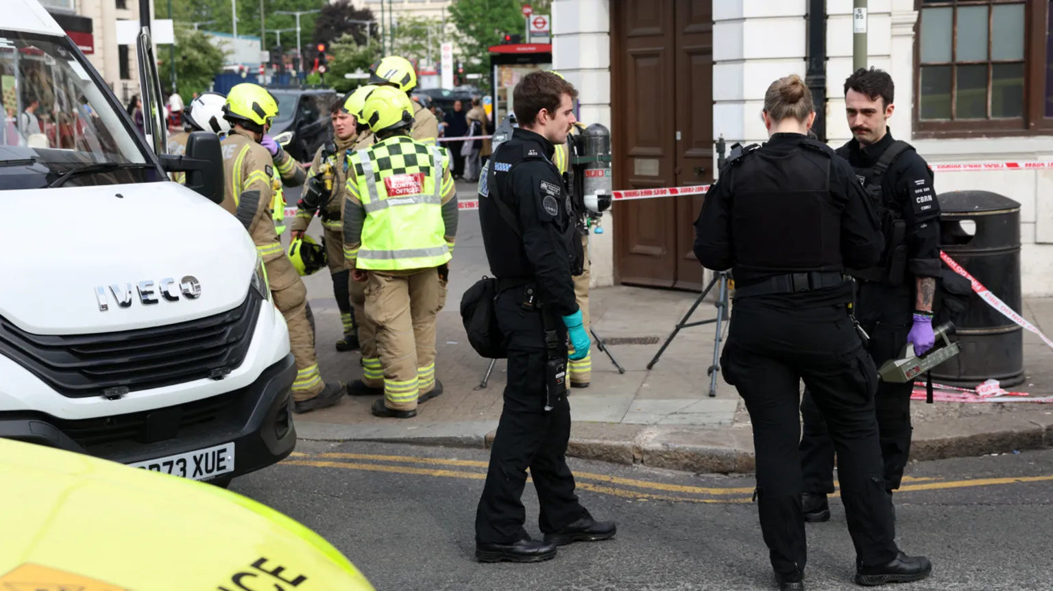  Police officers stood outside the corner of a bank which has been closed and there are also fire crews and a large white van parked close by, along with red and white tape blocking off the entrance to the bank, which is a light grey stone building with dark oak doors.