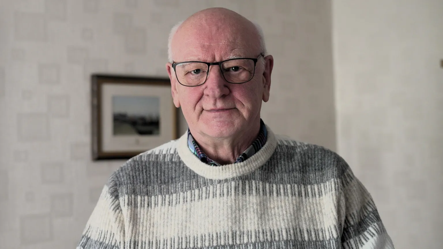 A balding man looks at the camera. It is a head and shoulders shot. He has black-rimmed glasses and is expressionless. He has a grey and white jumper on. Behind him are white walls with a framed photo on the wall.