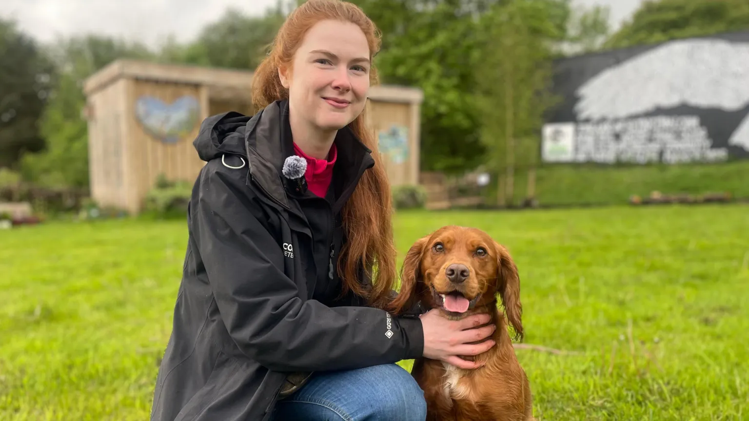 A woman crouches on one knee. She wears a black waterproof coat and a red jumper under it. She wears blue jeans and brown boots as she holds a dog, with both hands, that sits to her left. They pose on green grass with a wooden shed in the background. 