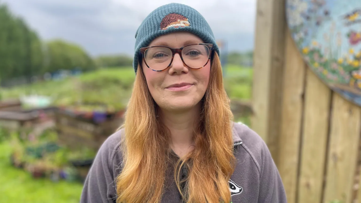A woman wears a blue beanie with an embroidered brown hedgehog on it. She has on clear, brown-rimmed glasses and has long red/blonde hair. Behind her, out-of-focus, is a garden with dozens of plants in it. 