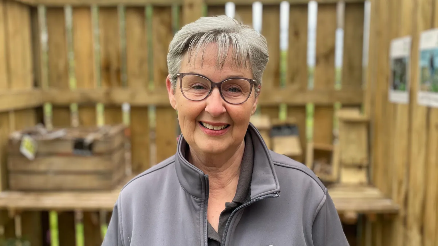 A woman has short grey hair and wears clear glasses with grey frames. She smiles at the camera as she stands inside a wooden hut wearing a grey zip-up jacket. 