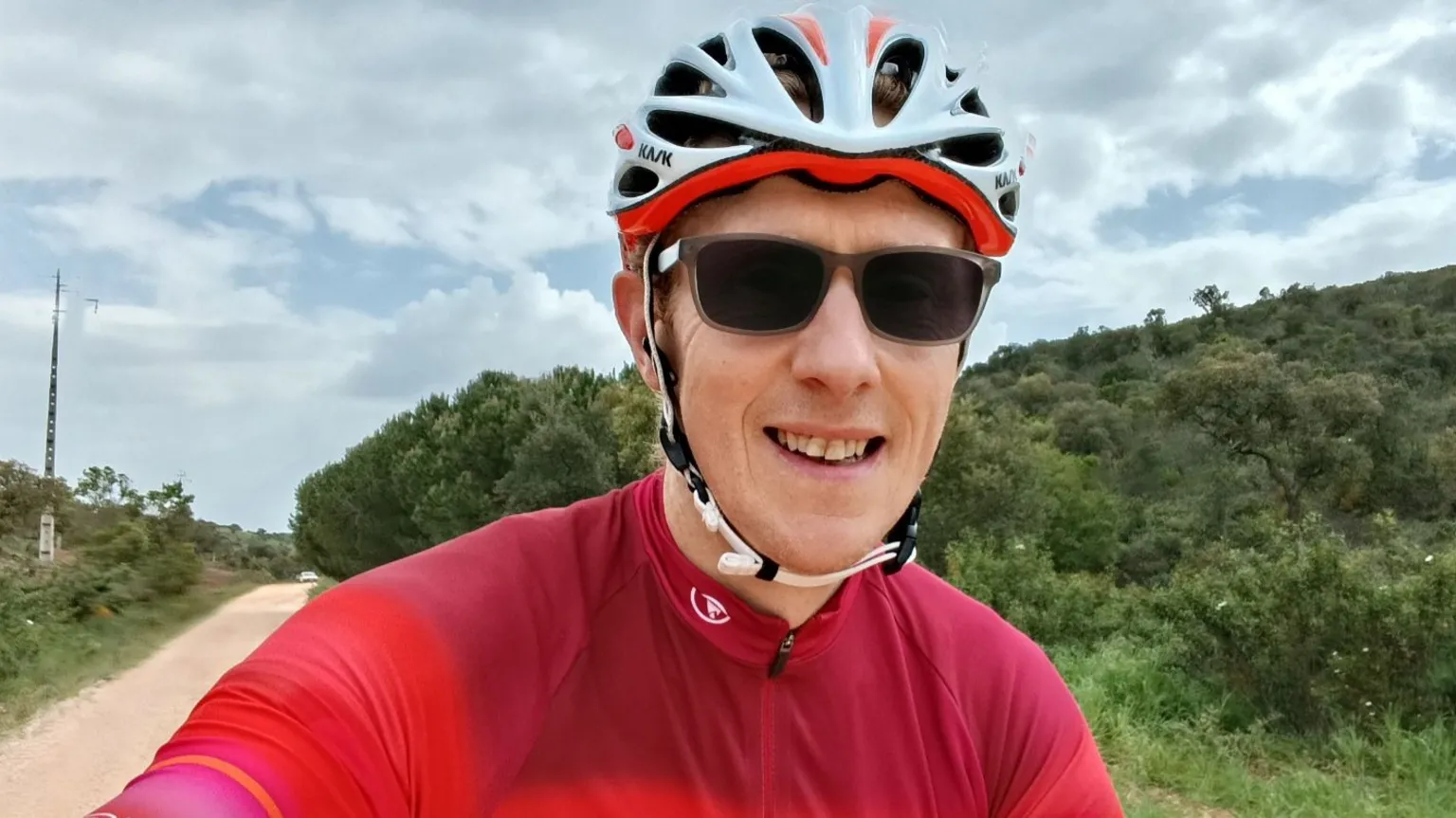 Victor Thompson A man wearing sunglasses, a bicycle helmet and red lycra smiles into the camera. He is outdoors and is cycling on a dusty road with green bushes and overgrowth on each side of the road.