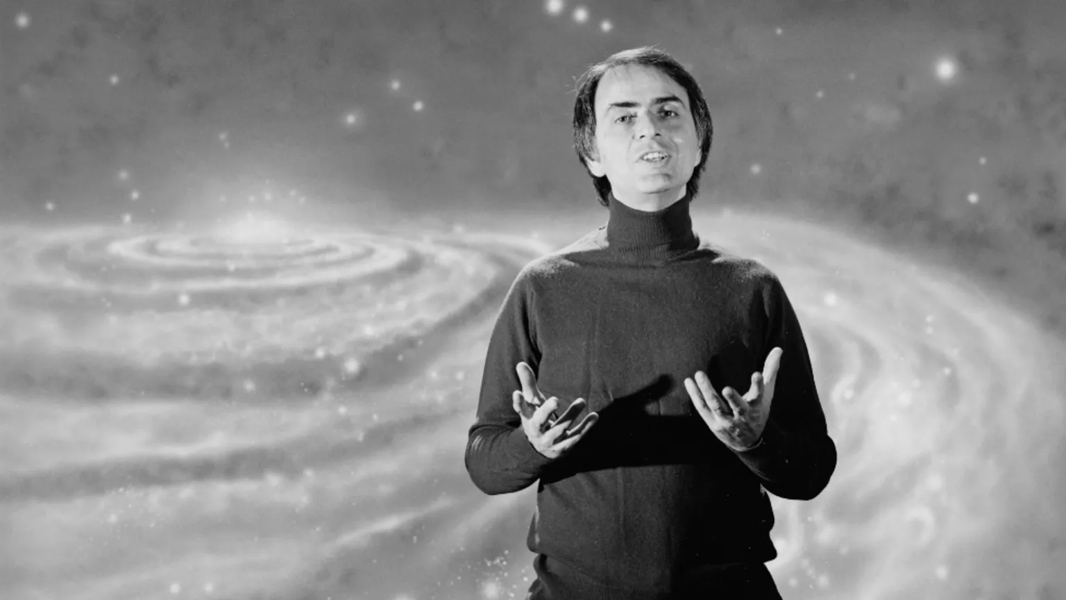  Black and white image of Carl Sagan, who has combed over dark hair and is wearing a turtle neck jumper. Behind him is a graphic of the universe.