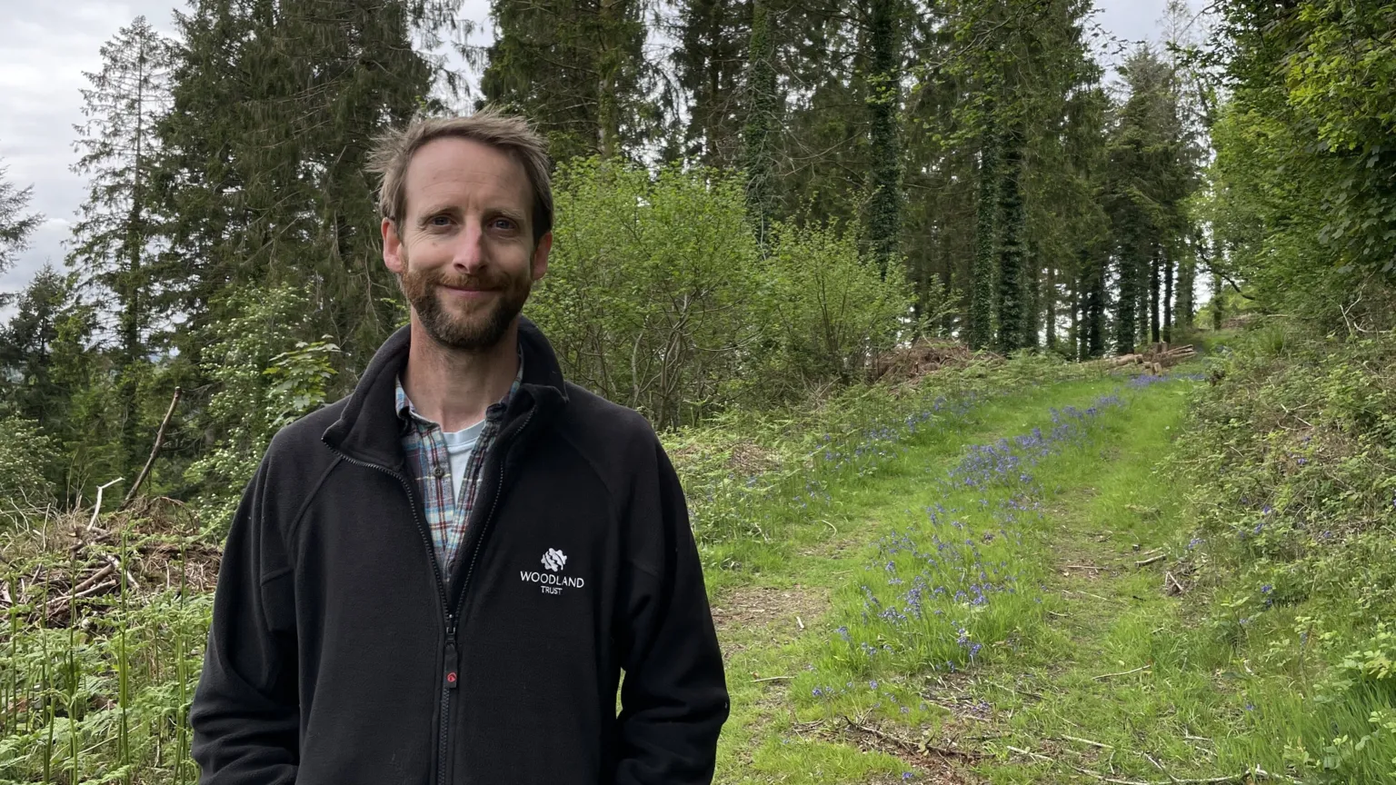 Joe Middleton wears a Woodland Trust fleece. He is standing in front of a path of bluebells stretching into the distance.