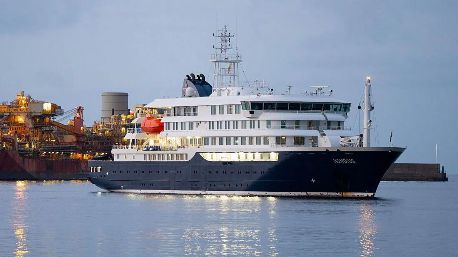 A medium-sized cruise ship, with the name Hondius visible, in port in early morning light, with other vessels behind it