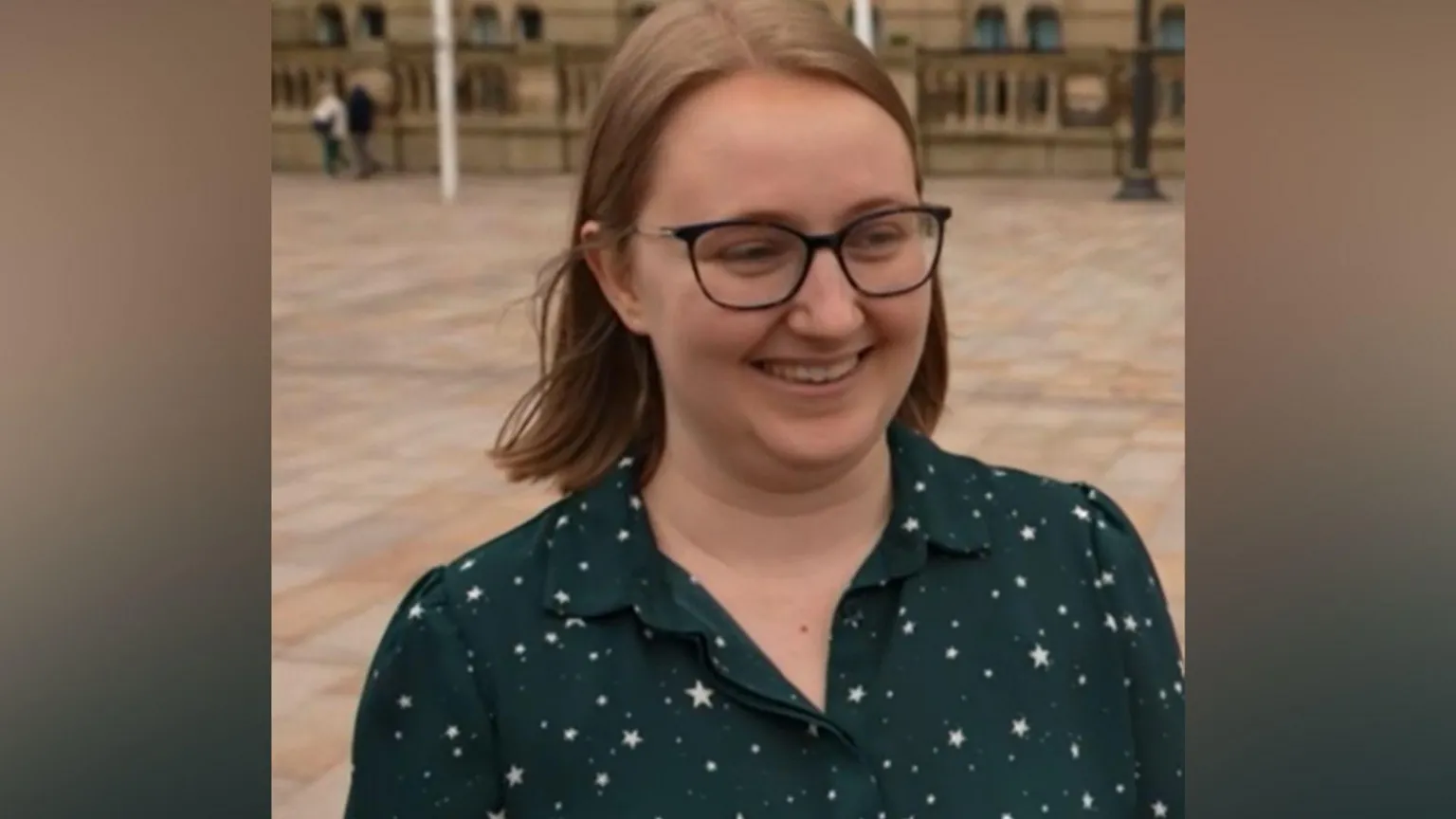 A woman with shoulder-length brown hair is pictured smiling while wearing thin framed black glasses. She is also wearing a dark green blouse with white stars dotted across it.
