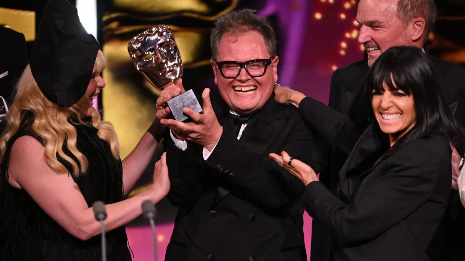  Alan Carr smiling and holding the Bafta trophy aloft, flanked by Paloma Faith and Claudia Winkleman