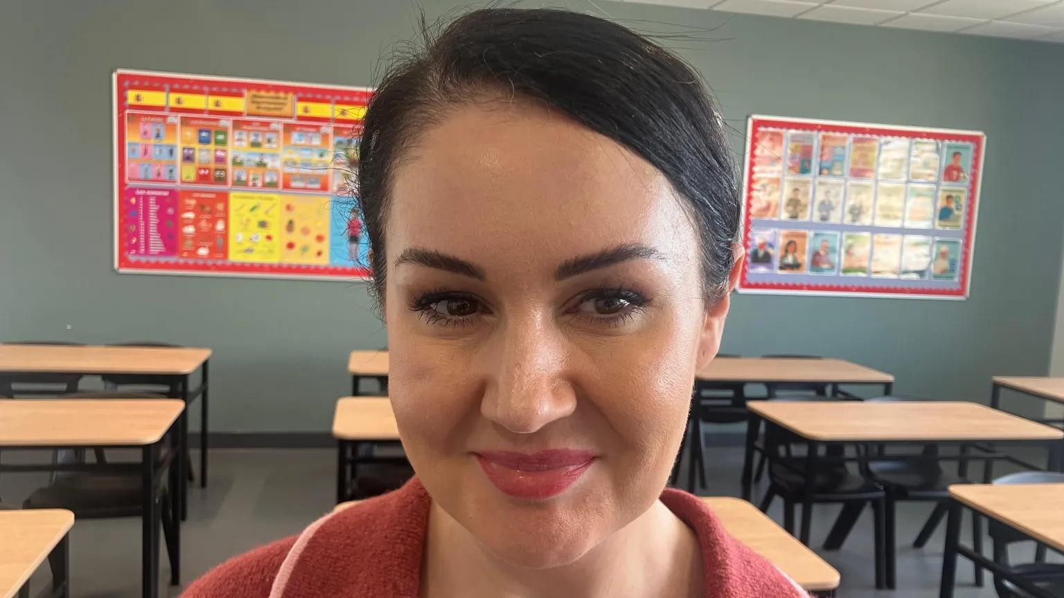 Olivia has black hair pulled back tight to the right. She is wearing a reddish v neck jumper with a loose collar.
