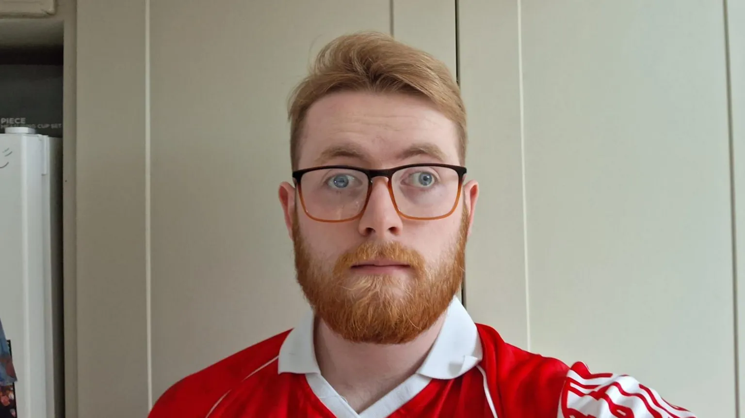 Cathal has ginger hair, lifted at the front, and a well-kept ginger beard. He wears glasses with square frames. His jersey is red with white stripes and a white collar.