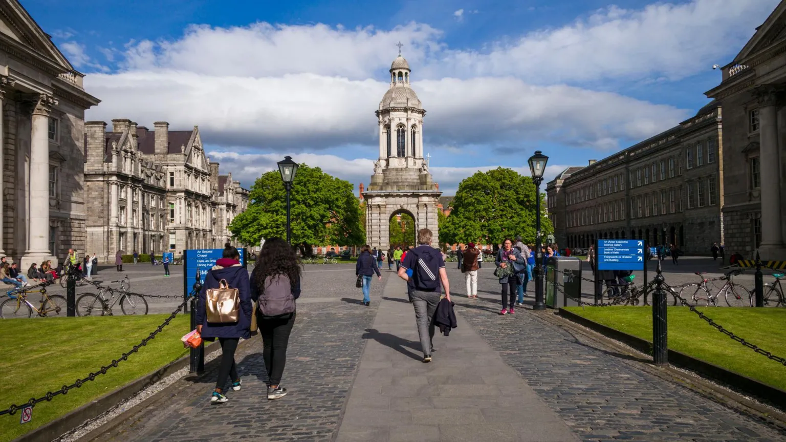 People walk with their backs to the camera through Trinity College Dublin's campus. The campus is paved and has some grass areas. It is surrounded by large old grey buildings. The centre of the campus has a large tower.