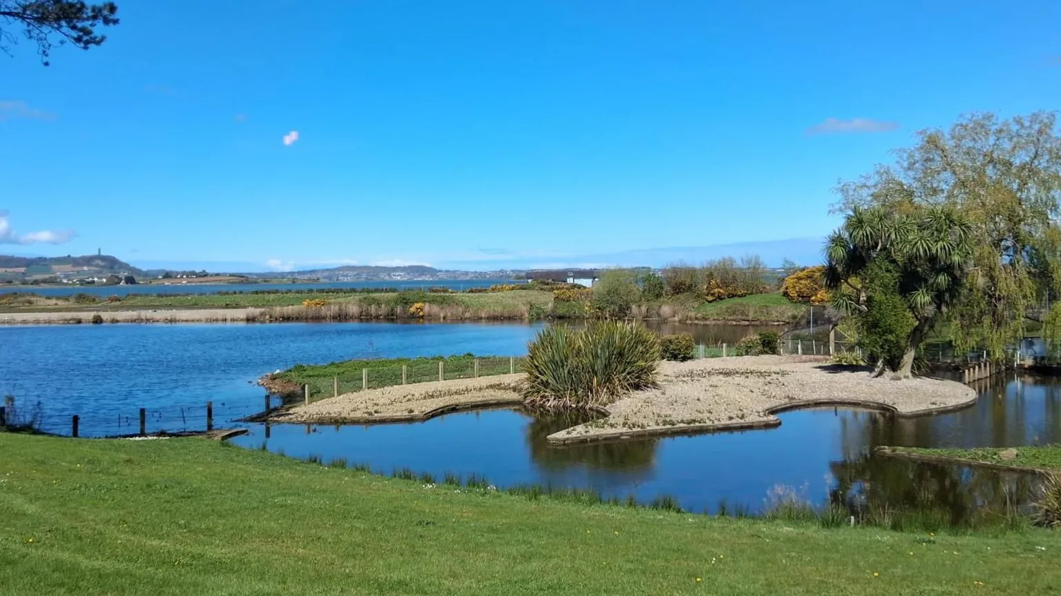 Crown Copyright Beyond a patch of kept green grass, blue water reaches towards a stretch of sand and more grass. The sky is clear, bar a few faint clouds. In the distance there are mountai9ns.