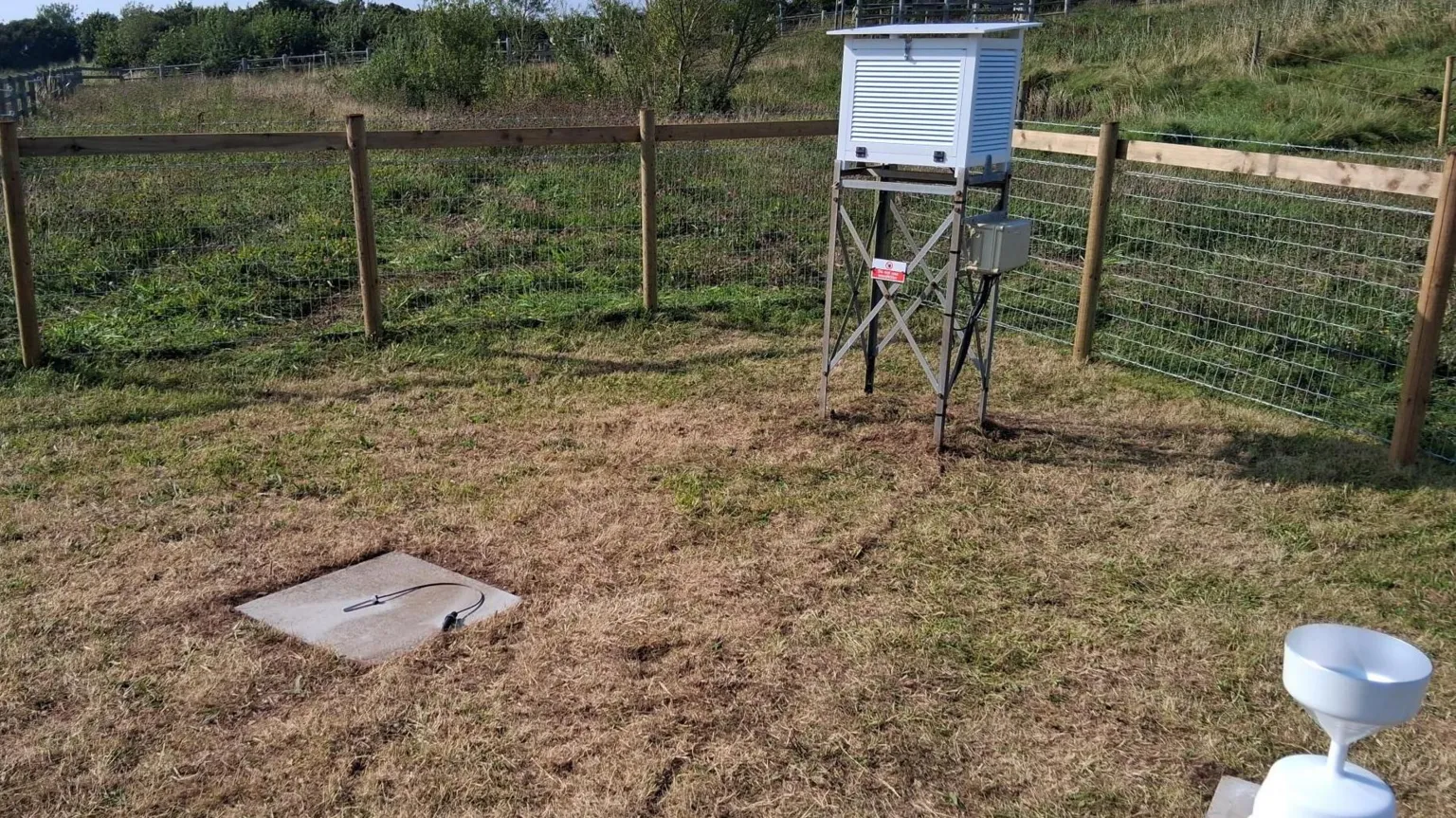 Crown Copyright Inside a small boundary, fenced off with wood and chicken wire, there is a wooden box propped on stilts, a concrete square on the ground, and a small white rain gauge