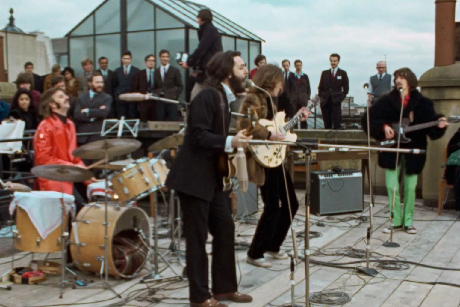 Apple Corps The Beatles play on the roof of 3 Savile Row. Paul McCartney is in the foreground, leaning into his microphone, while Ringo plays drums in a red leather jacket. Behind them are John Lennon and George Harrison, while dozens of suited men look on.