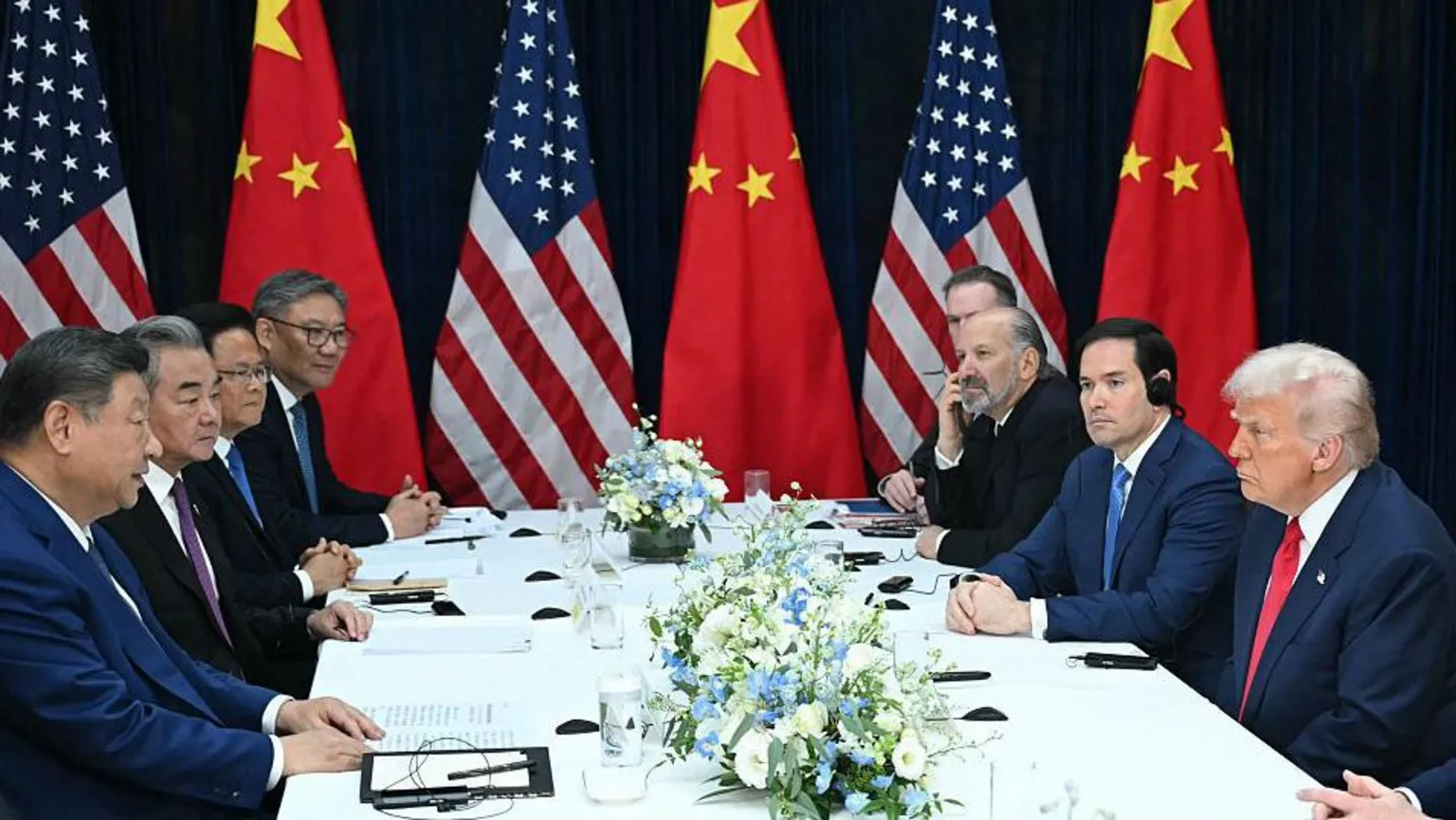AFP via President Xi facing Trump across a meeting table, flanked by their countries' representatives for bilateral talks. Flags of the US and China are displayed in the background.