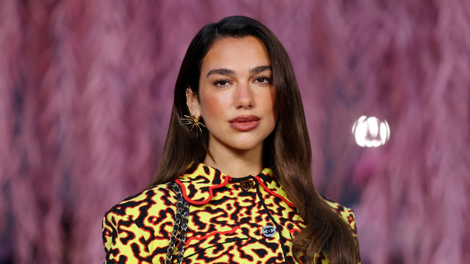 Dua Lipa, a young woman with long brown hair and a yellow and black patterned top, poses for the camera at a Chanel fashion show in Paris