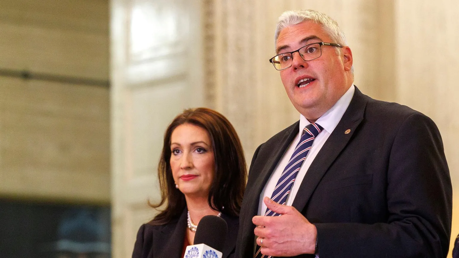 DUP Deputy First Minister Emma Little-Pengelly with party leader Gavin Robinson in the Great Hall at Stormont Great Hall. On the right - Gavin Robinson - a tall man with grey hair and glasses. He is speaking into a microphone and is wearing a suit and tie, On the left - Emma Little Pengelly has long dark brown hair, she is wearing a dark coloured jacket and a pearl necklace