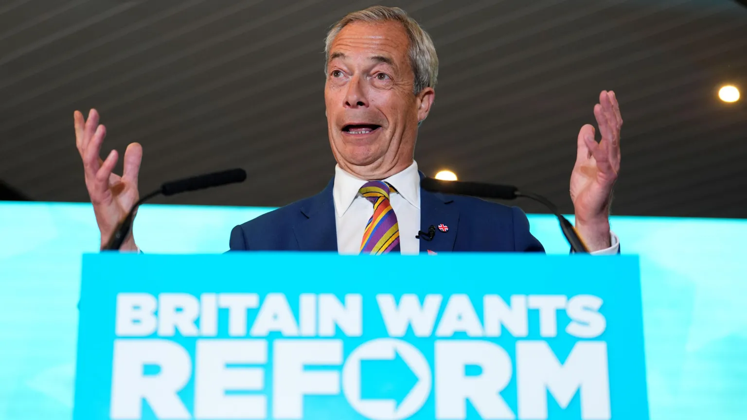  Nigel Farage has his hands raised while standing behind a podium, which is branded turquoise with words reading BRITAIN WANTS REFORM. He is wearing a navy blue suit and a striped multi-coloured tie.