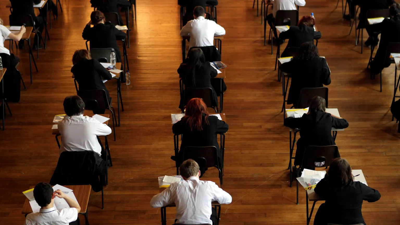  Rows of boys and girls in school uniform sitting at individual tables, appearing to be in the middle of an exam. They are photographed from the back