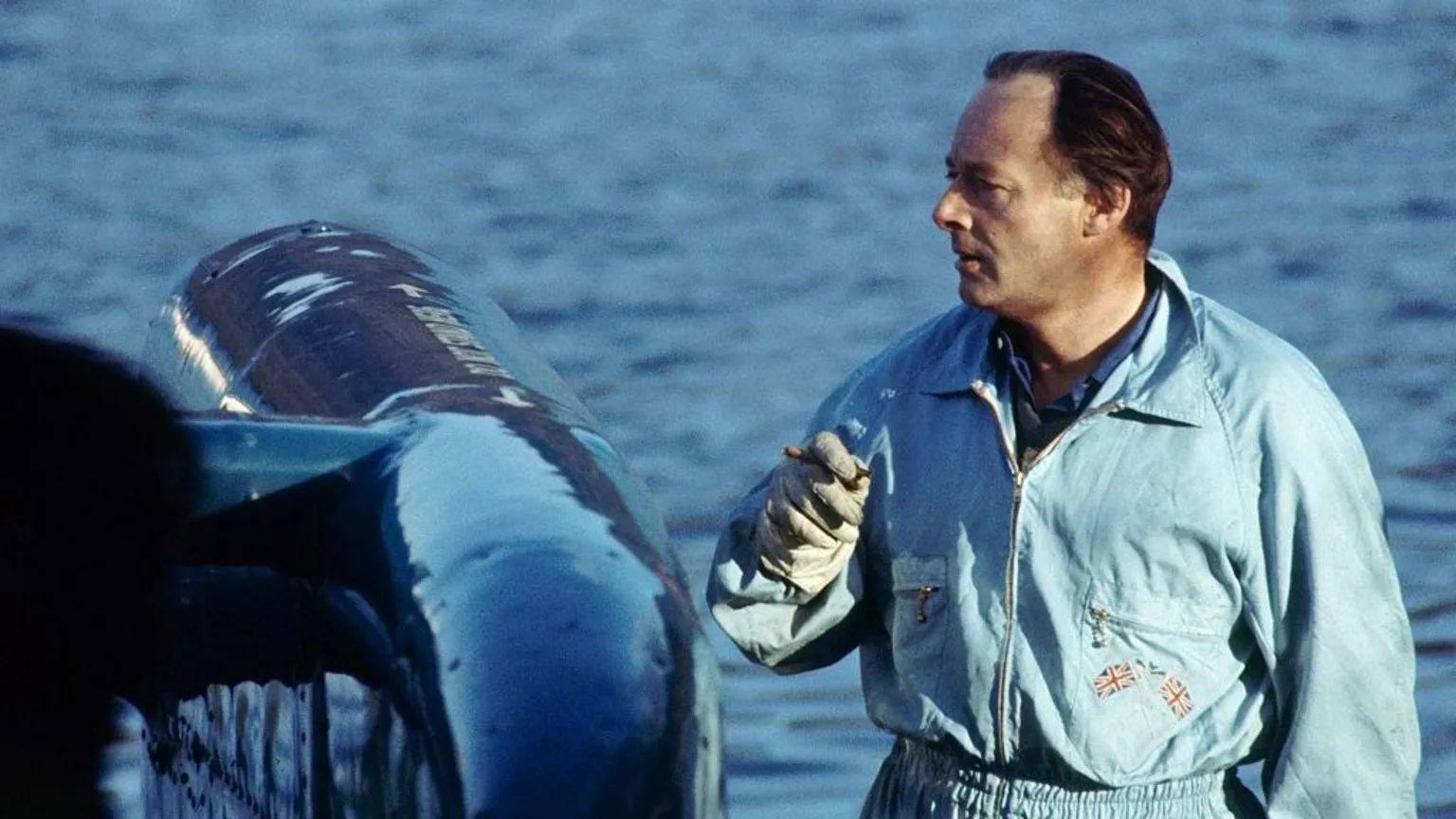  Donald Campbell smokes a cigar as he stands next to Bluebird in January 1967. He has dark hair brushed back from his face and is wearing blue overalls.