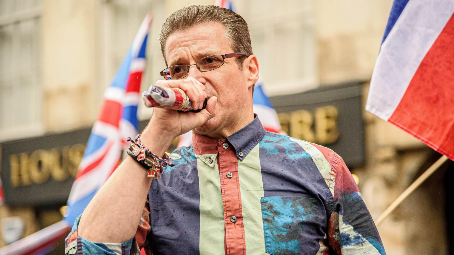  Alistair McConnachie, who has short brown hair, speaks into a microphone. He is wearing a union flag coloured shirt. 