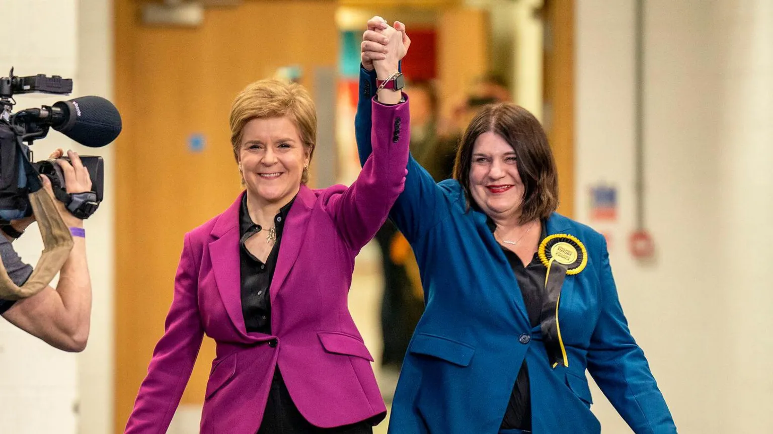  Nicola Sturgeon and Susan Aitken hold hands lifted into the air in a show of triumph. Sturgeon has short fair hair and wears a pink blazer with a black shirt, and Aikten has shoulder-length dark brown hair and wears a blue blazer, black shirt, and yellow and black SNP rosette. A TV camera films them