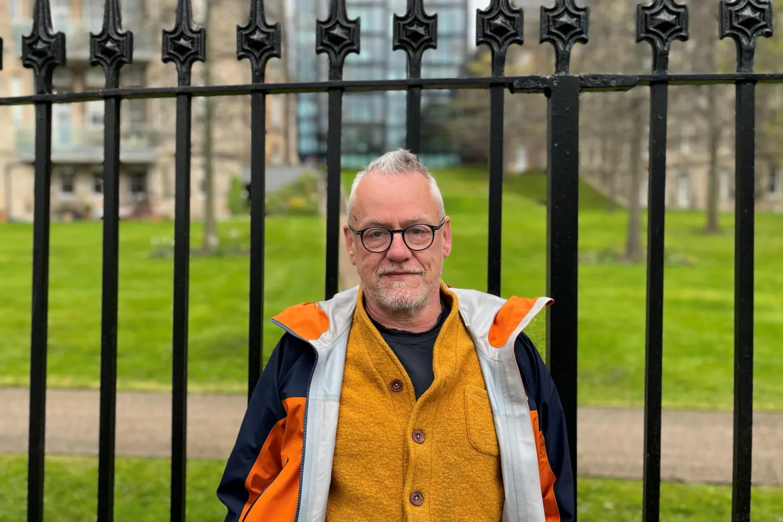 A man with grey hair and glasses stands in front of black railings wearing a yellow cardigan and orange jacket