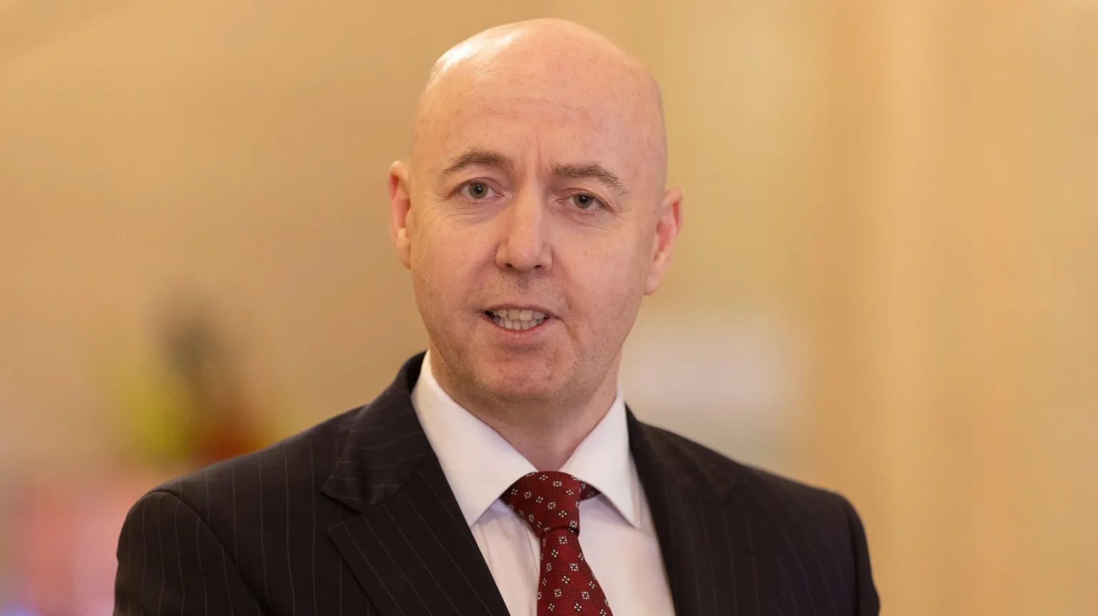 A bald man wearing a dark pinstriped suit, white shirt an red tie.