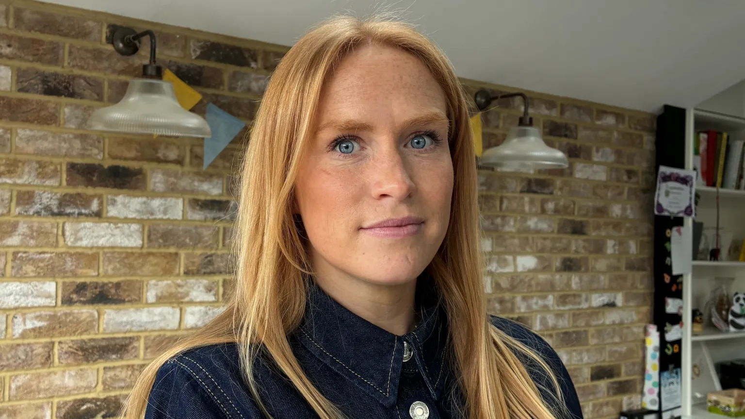 Close-up portrait of a woman with long light hair and light-coloured eyes, wearing a dark denim shirt, standing indoors in front of a light brick wall with two wall-mounted lamps and shelves visible in the background.