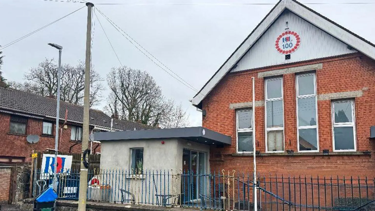 The image is of a number of buildings, including the Antrim branch of the Royal British Legion. It's pictured on the right and is a red brick building with tall windows and a Royal British Legion 100 emblem. It's surrounded by smaller red brick buildings and a blue metal fence. 